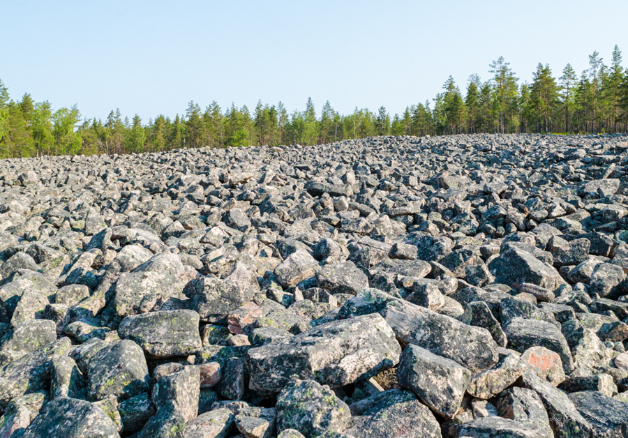 Suuri kivipelto kuvattuna lähietäisyydeltä. Etualalla kiviä, horisontissa näkyy metsää sekä pilvetön taivas.