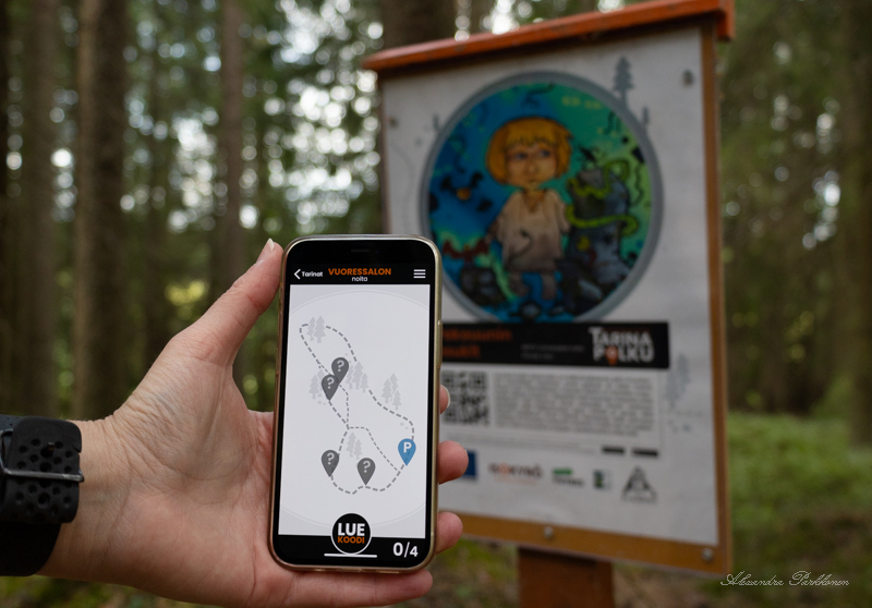 Kuva, jossa näkyy Tarinapolku-sovellus puhelimen näytöllä sekä sen takana metsässä oleva Tarinapolku-sovelluksen opastekyltti.