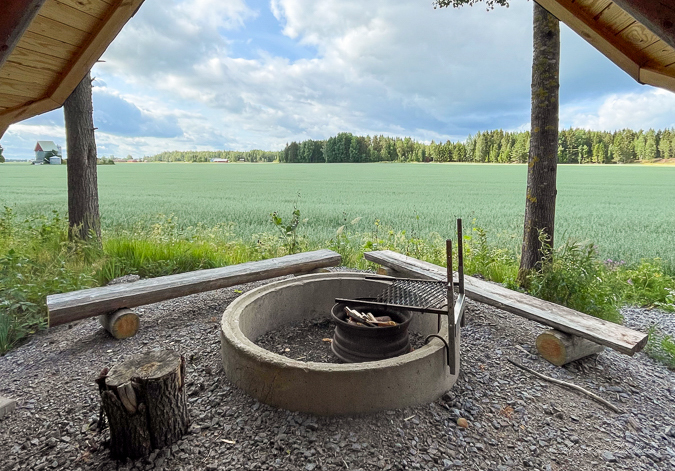 Laavun nuotiopaikalta otettu kuva, jossa näkyy maisema joka avautuu laavulta. Maisemassa näkyy nuotiopaikan ja penkkien lisäksi peltoa ja metsää.