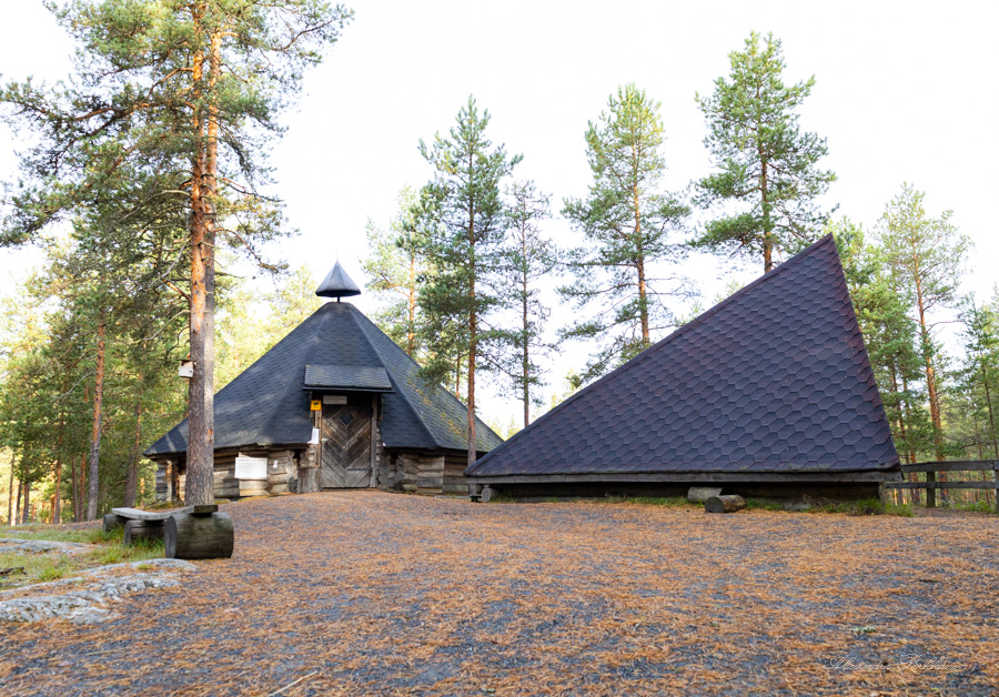 kuva metsässä sijaitsevasta kodasta. Etualalla näkyy kivituhkapintaista maata, jossa on neulasia. Kodan ympärillä on puita.