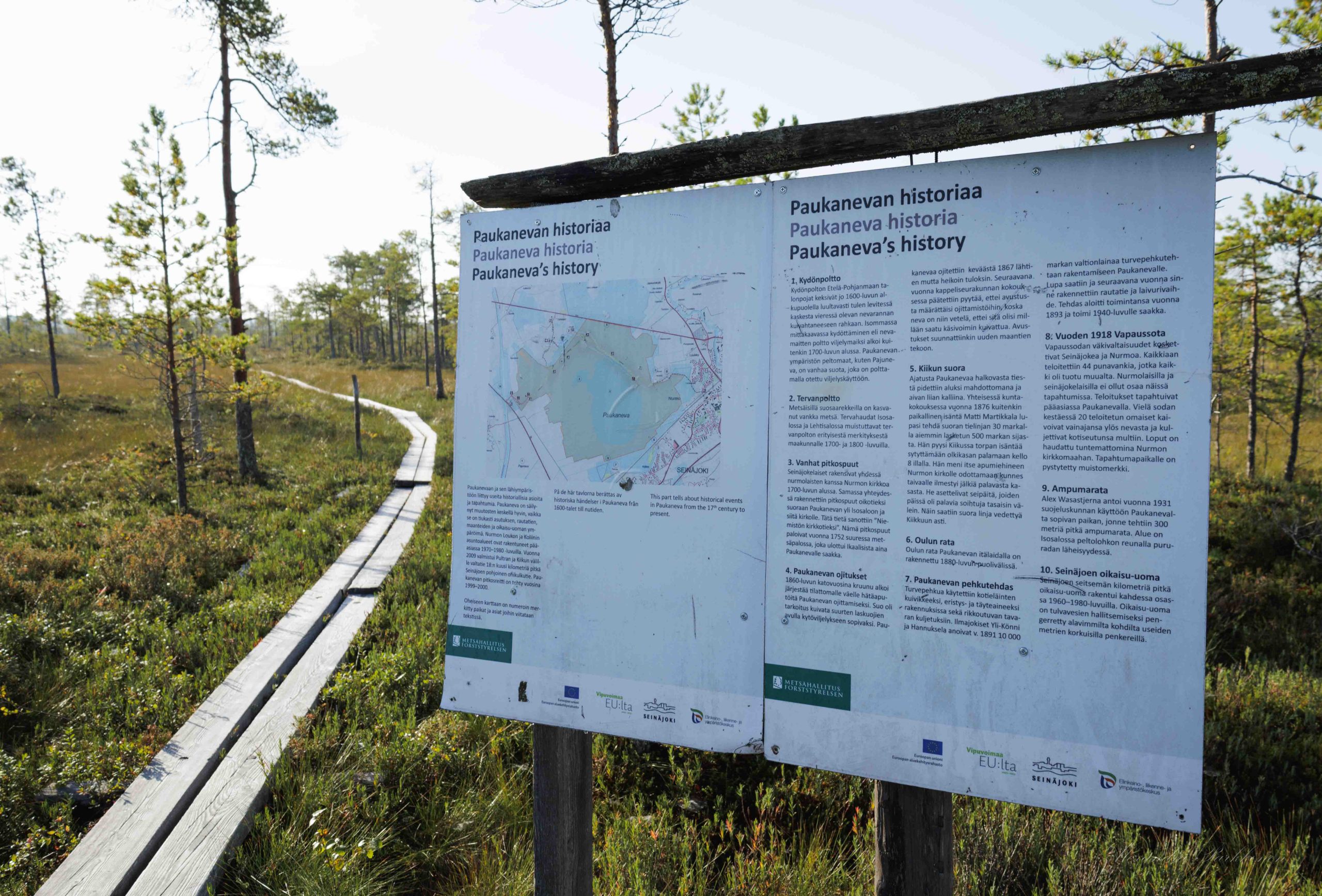 Lähikuva retkeilyreitin opastetaulusta. Taustalla näkyy pitkospuut