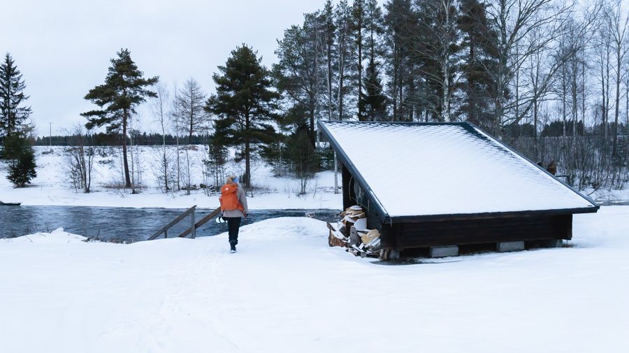Retkeilijä kävelee talvisessa maisemassa kohti laavua, joka sijaitsee joen rannassa.