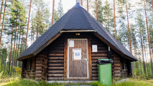 Lähikuva hirsisestä kodasta, jonka edustalla on vihreä roskapönttö. Kodan takana näkyy metsää.