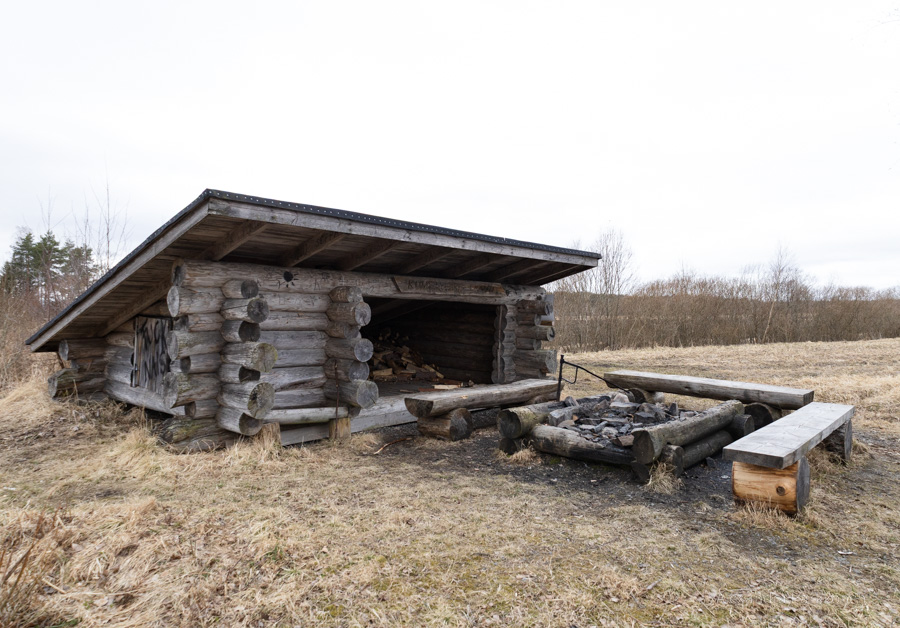 Puinen laavu kuvattuna etuviistosta. Laavun edustalla on puisin penkein reunustettu nuotiopaikka. Etualalla näkyy nurmikkoa.