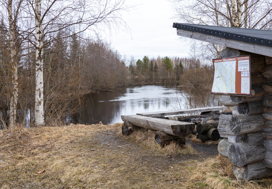 Maisemakuva laavulta, jossa on näkymä joelle. Oikealla näkyy laavua, jonka edustalla on puisin penkein reunustettu nuotiopaikka. Kauempana näkyy joki sekä metsää.