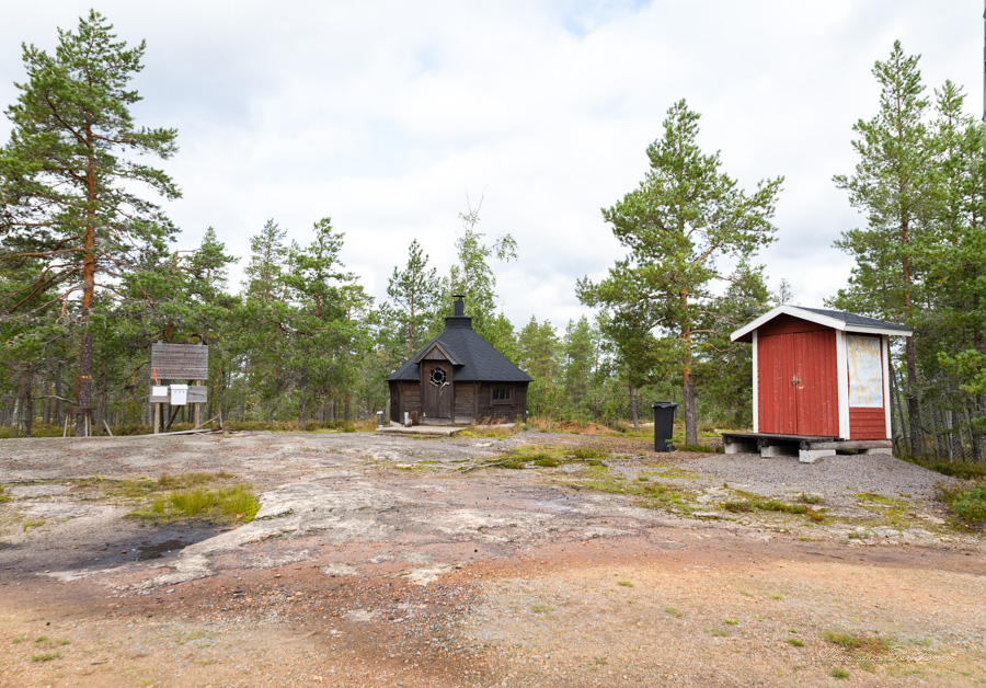 Kota, jonka vieressä on punainen puuvaja. Etualalla näkyy kalliota ja kodan takana metsää.