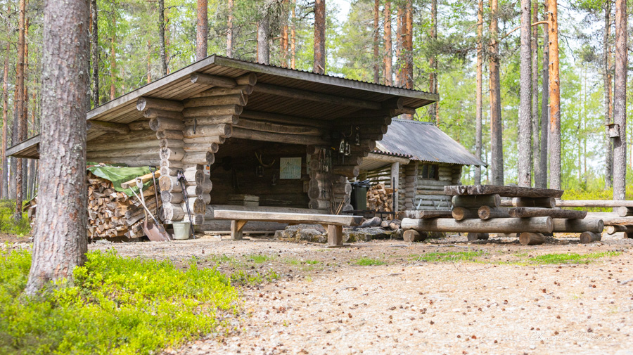 Laavu kuvattuna etuviistosta. Laavun edustalla on tulipaikka ja puinen pöytä penkkeineen. Ympärillä on mäntymetsää.